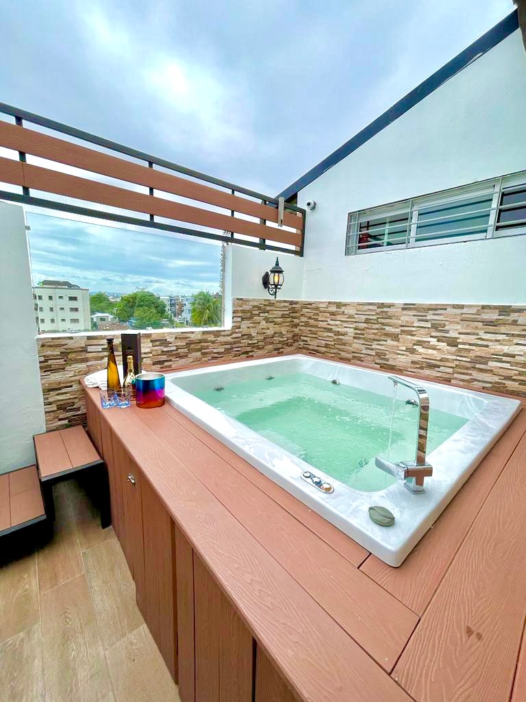 A relaxing hot tub is situated on a wooden deck, surrounded by a stone accent wall and a railing. A scenic view is visible in the background, complementing the serene environment. Bottles and colorful glasses are placed on the deck, enhancing the inviting space.