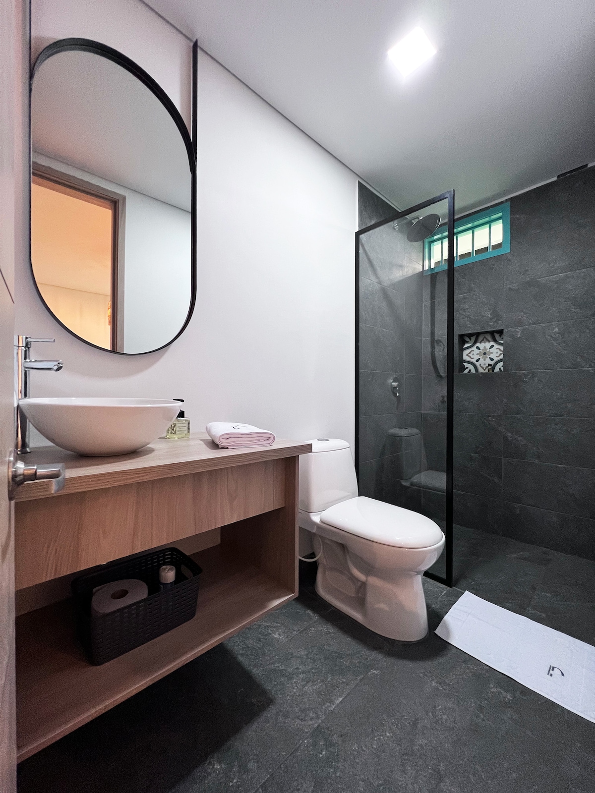 A modern bathroom is featured, showcasing a glass shower enclosure, a sleek vessel sink on a wooden vanity, and a circular mirror. Neutral tiles create a cohesive design, and a small basket is visible containing toiletries. A white bath mat adds a touch of softness to the space.