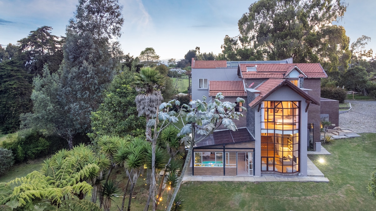 The exterior of a spacious mansion features a modern design with large glass windows and a prominent rooftop. Surrounding greenery includes various trees and shrubs, providing a natural setting. The building's structure showcases multiple levels and a welcoming entrance, complemented by well-maintained outdoor areas.