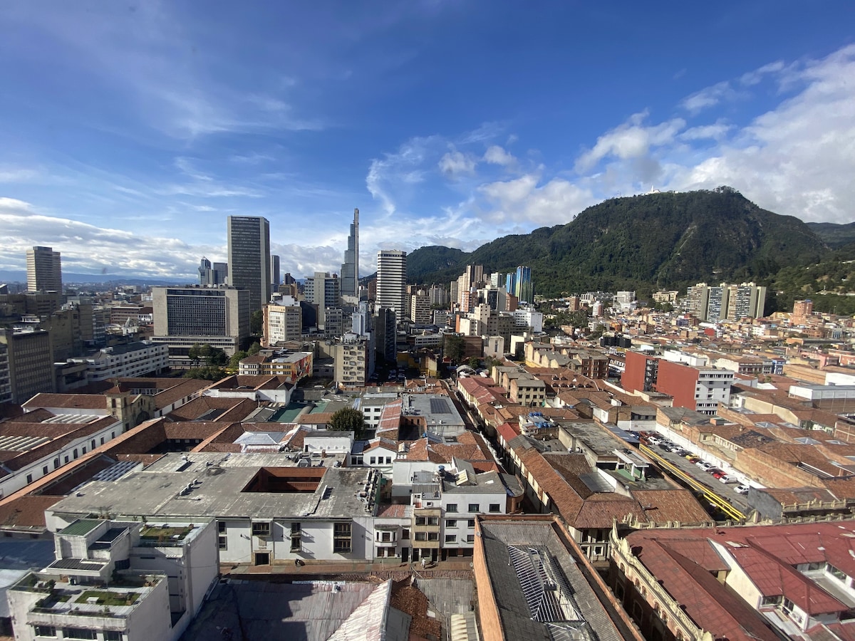 A panoramic view of Bogotá reveals a mix of modern buildings and traditional rooftops. The skyline is dominated by tall structures, while the presence of green mountains in the background adds depth to the urban landscape. A range of colors highlights the diverse architecture.