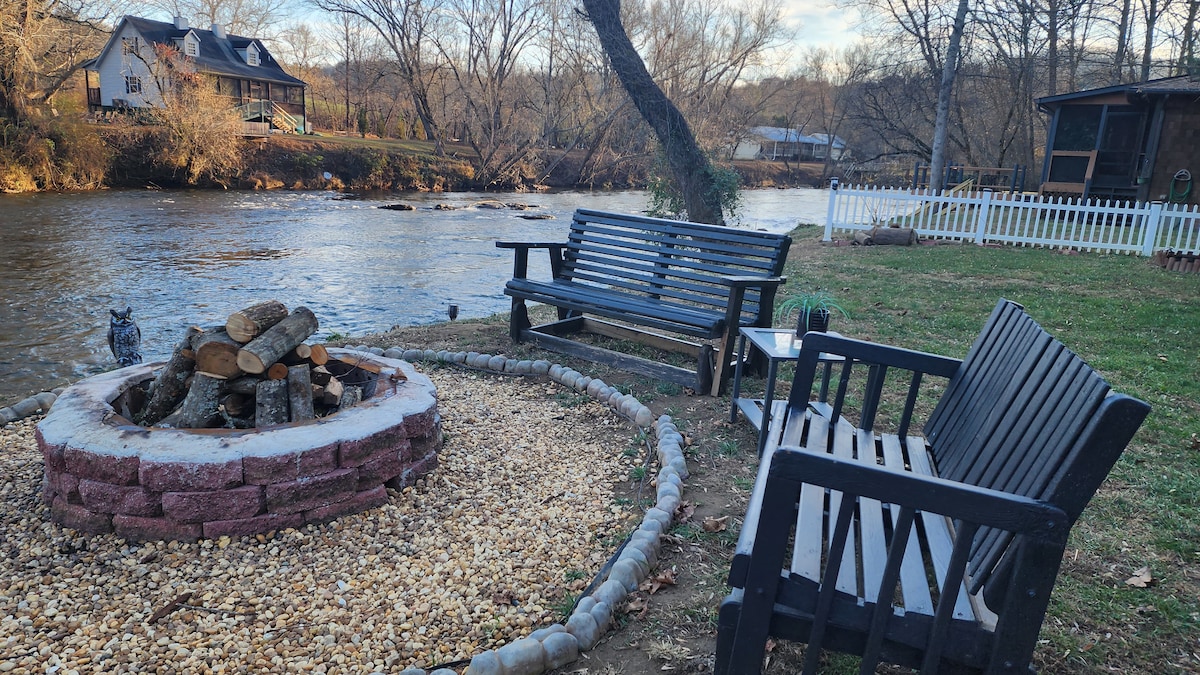 Two black wooden benches are positioned beside a circular firepit made of stone, surrounded by small gravel. The firepit is adorned with logs. A serene view of the river and trees is visible in the background.