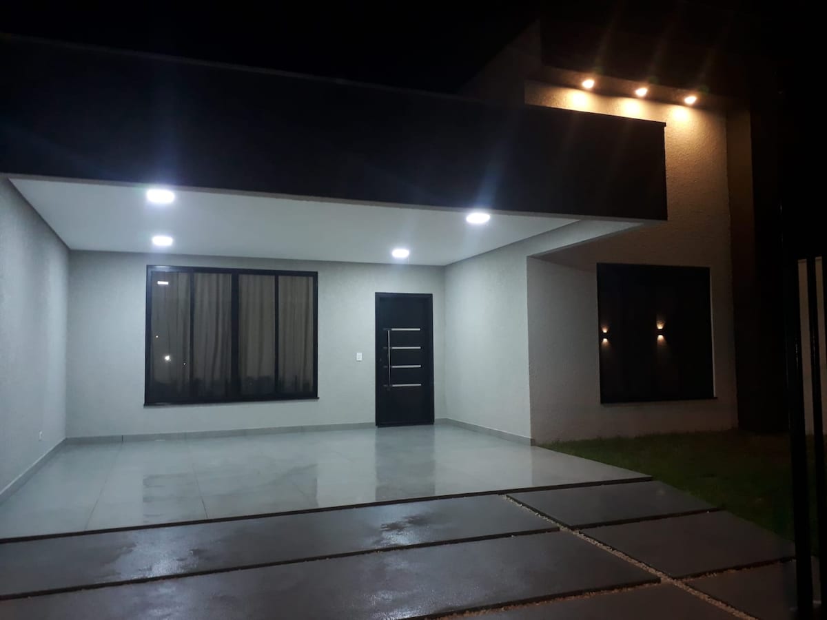 The exterior of the modern residence is showcased at night, featuring a polished concrete patio illuminated by soft overhead lights. Large windows reflect the light, and a sleek main entrance door stands prominently, contributing to the contemporary design.