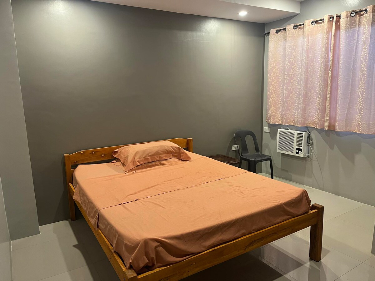A spacious sleeping area features a wooden bed with an orange bedspread and a matching pillow. A single chair is positioned beside the bed, and an air conditioning unit is visible on the wall, near a window with light floral curtains.