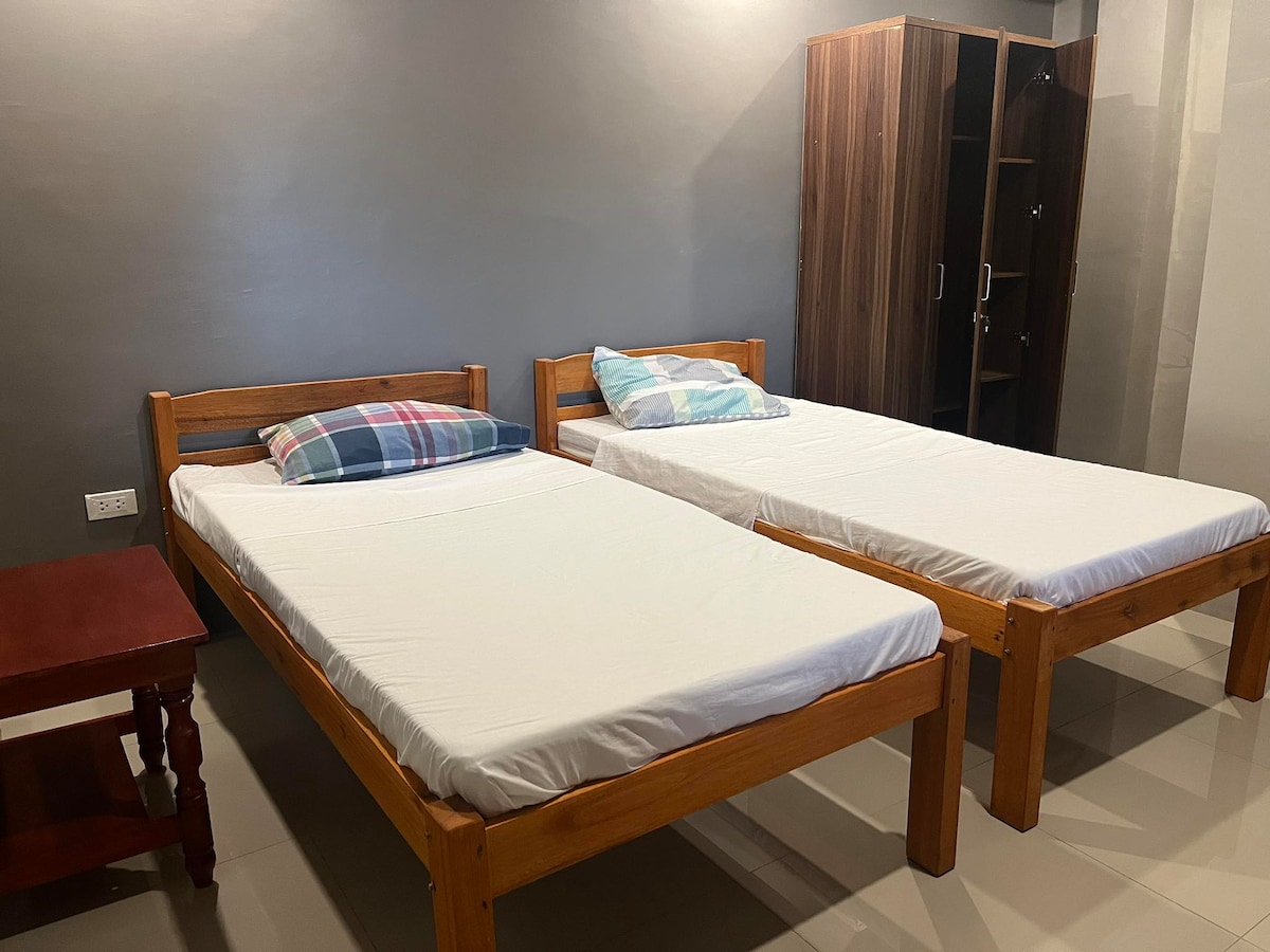 The room features two wooden twin beds with simple white bedding, complemented by colorful pillows. A small wooden table stands beside one bed, and a wardrobe is visible in the background, providing storage options.