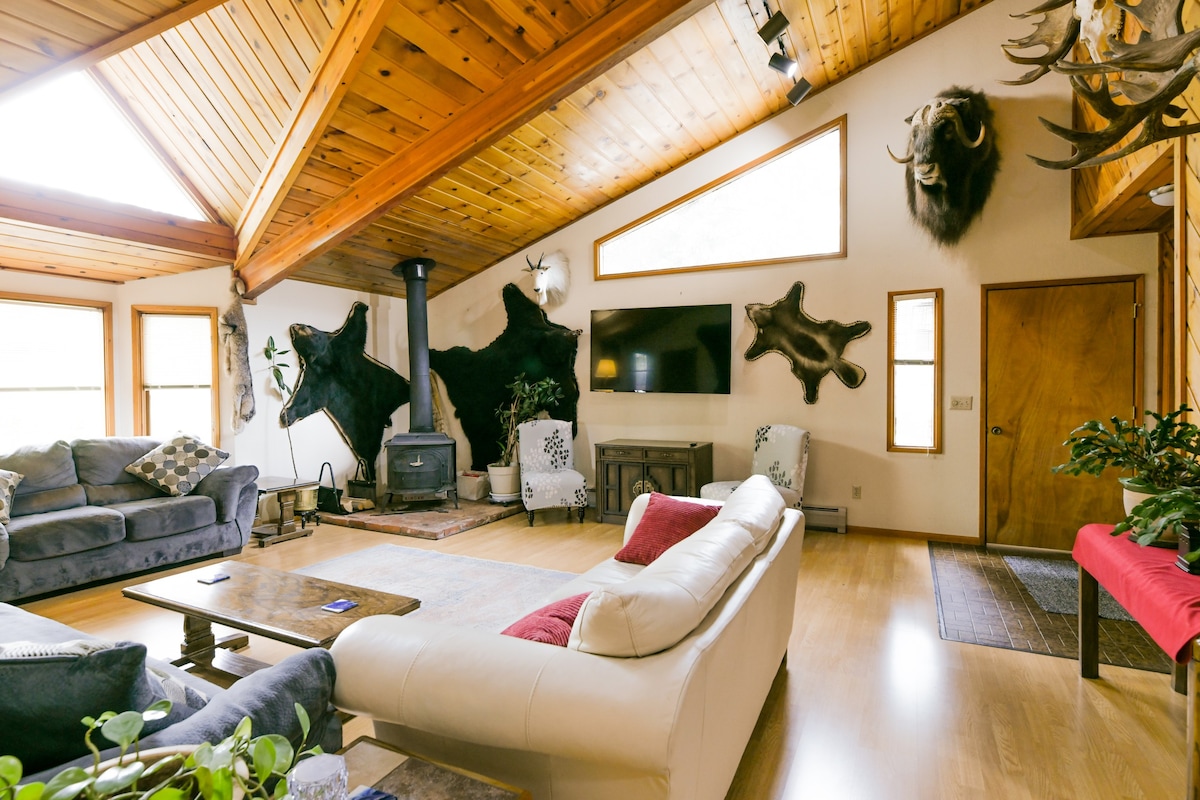 A spacious living room features a wooden ceiling with skylights, enhancing natural light. Comfortable seating arrangements include a white and gray sofa. Natural elements are present, with mounted animal heads on the walls and greenery in the room. A cozy fireplace and television are central to the space.