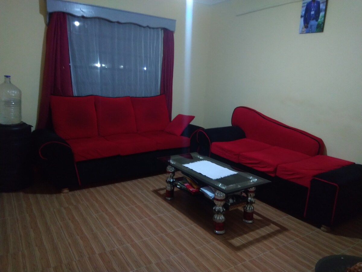 A living area is furnished with a black sofa set featuring red cushions, arranged around a glass coffee table. The room is highlighted by a window dressed with sheer curtains, while the warm-toned flooring complements the overall design.