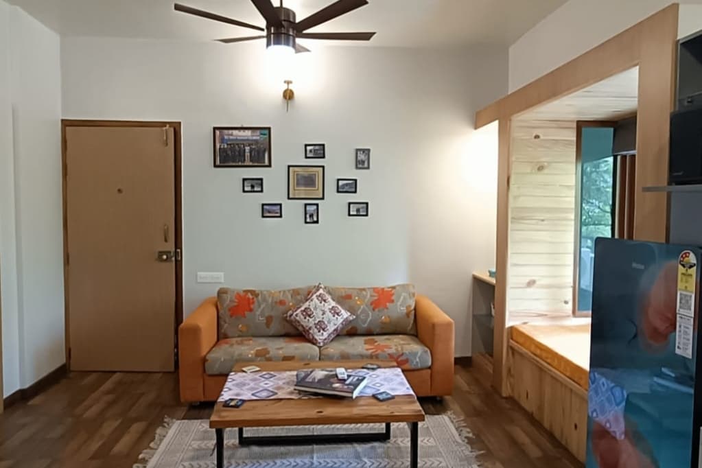 A comfortable living area features a sofa with bright orange and floral patterns, alongside a wooden coffee table. The walls display framed photographs, and warm light is provided by a ceiling fan above. A cozy reading nook is visible in the corner.