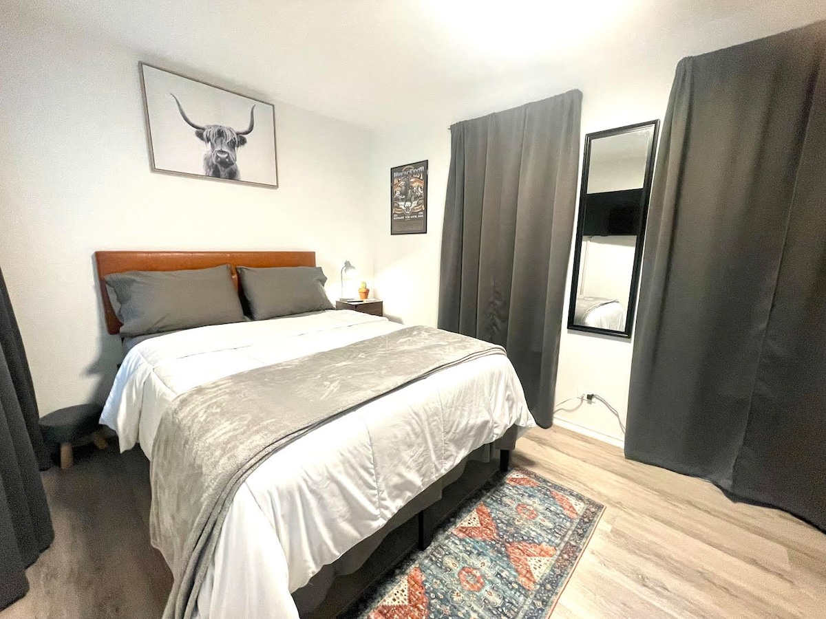 A bright sleeping area is furnished with a spacious bed dressed in soft linens. Dark curtains frame the window, allowing for privacy. A decorative rug adds warmth to the wooden flooring. A full-length mirror is positioned near the corner, enhancing the room's functionality.