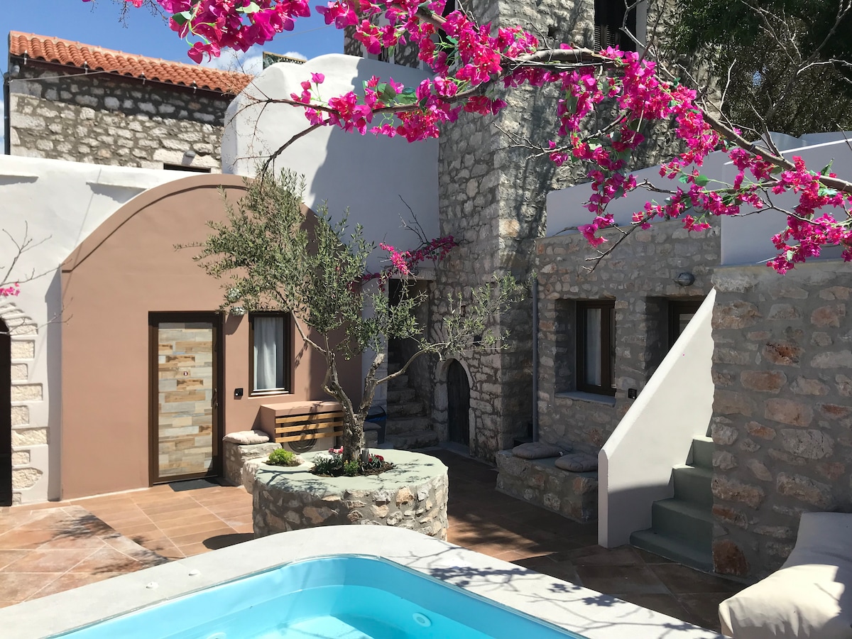 A charming outdoor area features a small swimming pool surrounded by stone walls. Vibrant pink bougainvillea branches extend across the scene, enhancing the rustic aesthetic. A path leads to an arched entryway and several windows that provide light to the space.