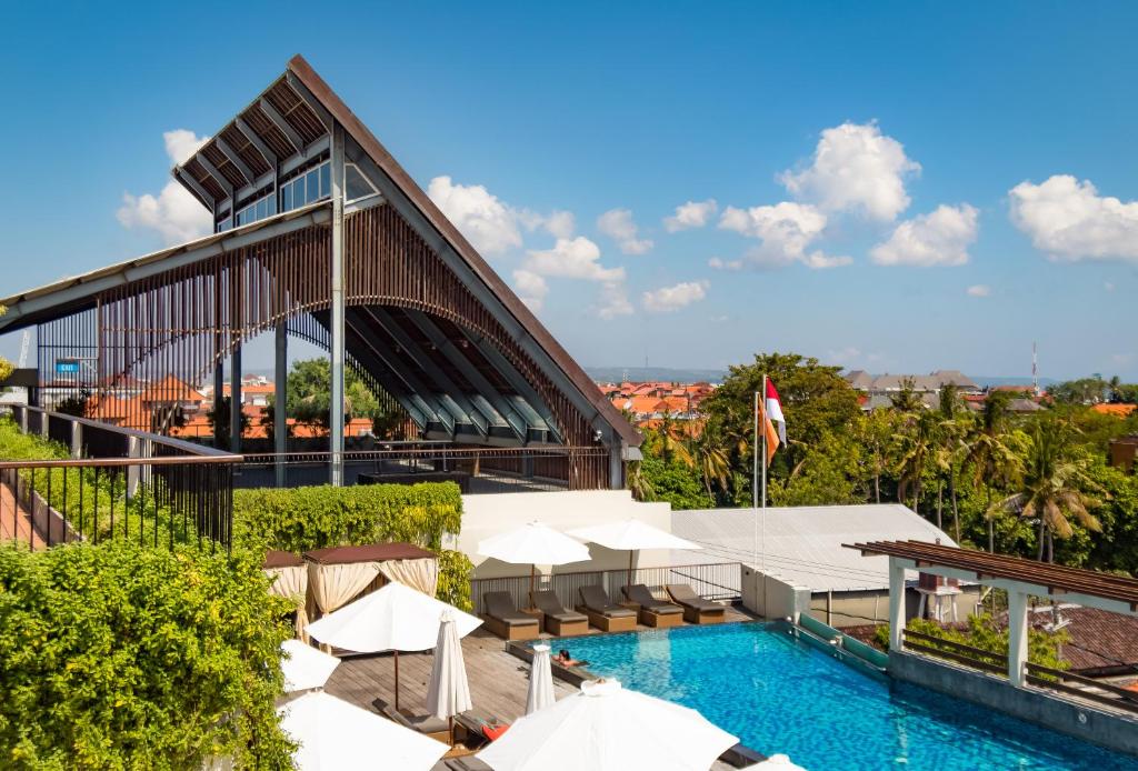 An outdoor pool area features sun loungers surrounded by greenery. An architecturally striking structure with a distinctive roof design overlooks the pool. The clear blue water reflects the sky, while shaded areas provide relaxation options. This space offers a blend of comfort and modern design.