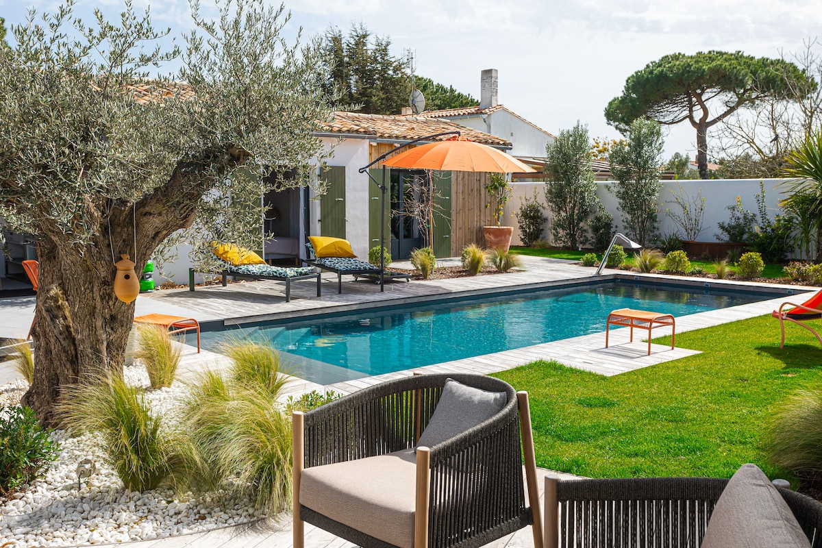 A tranquil outdoor area features a serene salt-water pool surrounded by lush greenery and artistic landscaping. Stylish seating arrangements are positioned near the pool, with vibrant orange umbrellas offering shade. An olive tree adds a natural element to the aesthetic harmony of the space.