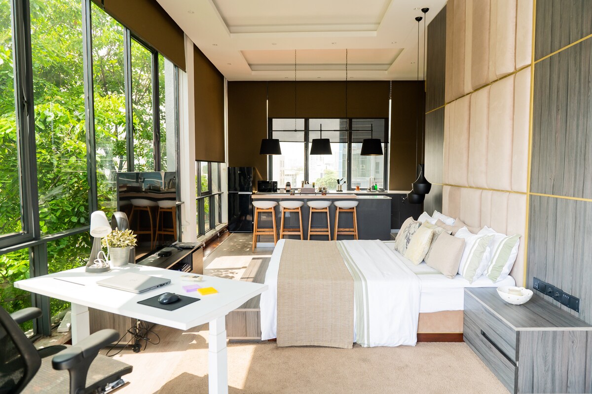 An open layout features a spacious bed with neutral bedding and a light throw. A designated workspace is positioned next to the bed, accompanied by a comfortable chair. Large windows fill the room with natural light, while modern pendant lights hang from the ceiling above a minimalist kitchen area.