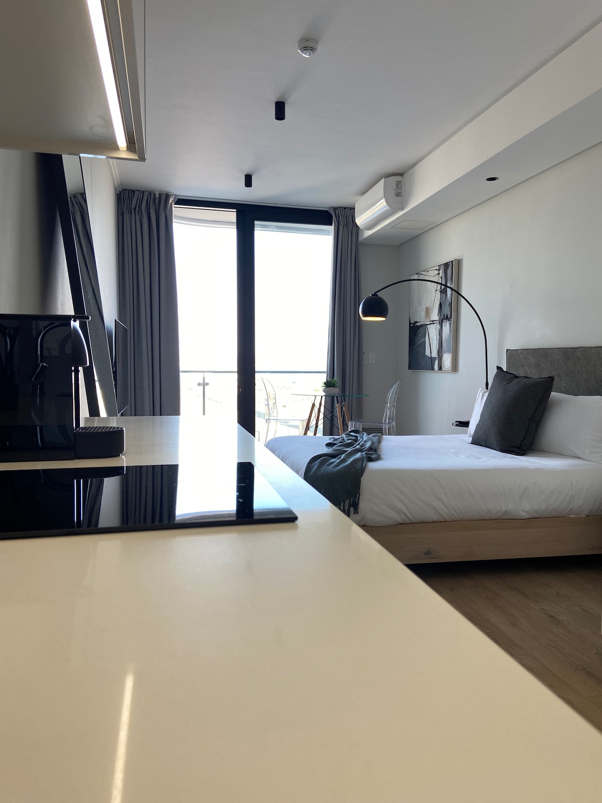 The studio apartment features a spacious bed with neutral bedding, complemented by accent pillows. A well-appointed kitchenette is visible in the foreground, along with sleek countertops. Floor-to-ceiling windows allow natural light to fill the space, offering views of a sunny balcony.