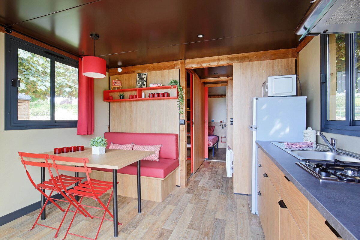 The interior space is designed with wood accents and features a dining area with a wooden table and red chairs. A cozy seating nook is visible along one wall, and the kitchenette is equipped with a stove and microwave. Two windows provide natural light.