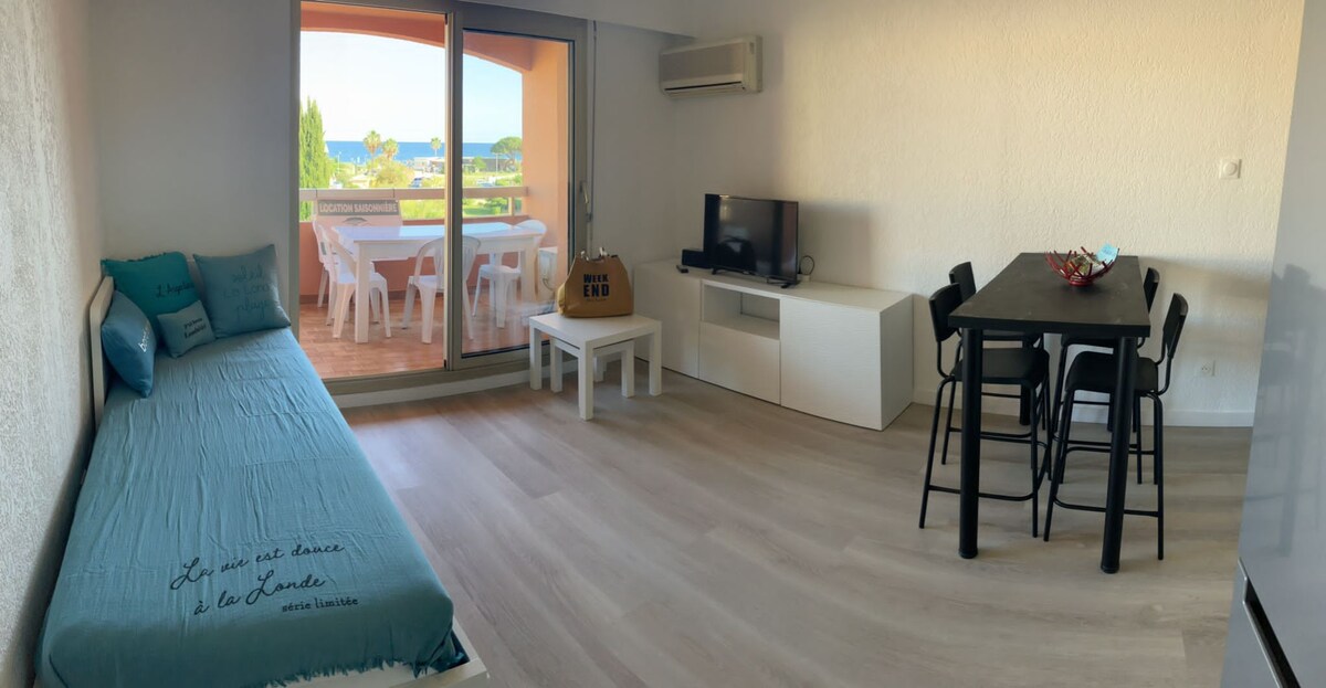 A bright living area features a comfortable sofa bed and a dining table surrounded by black chairs. Light wood flooring complements the minimalist decor. Sliding glass doors open to a terrace with a sea view, providing an inviting connection to the outdoors.