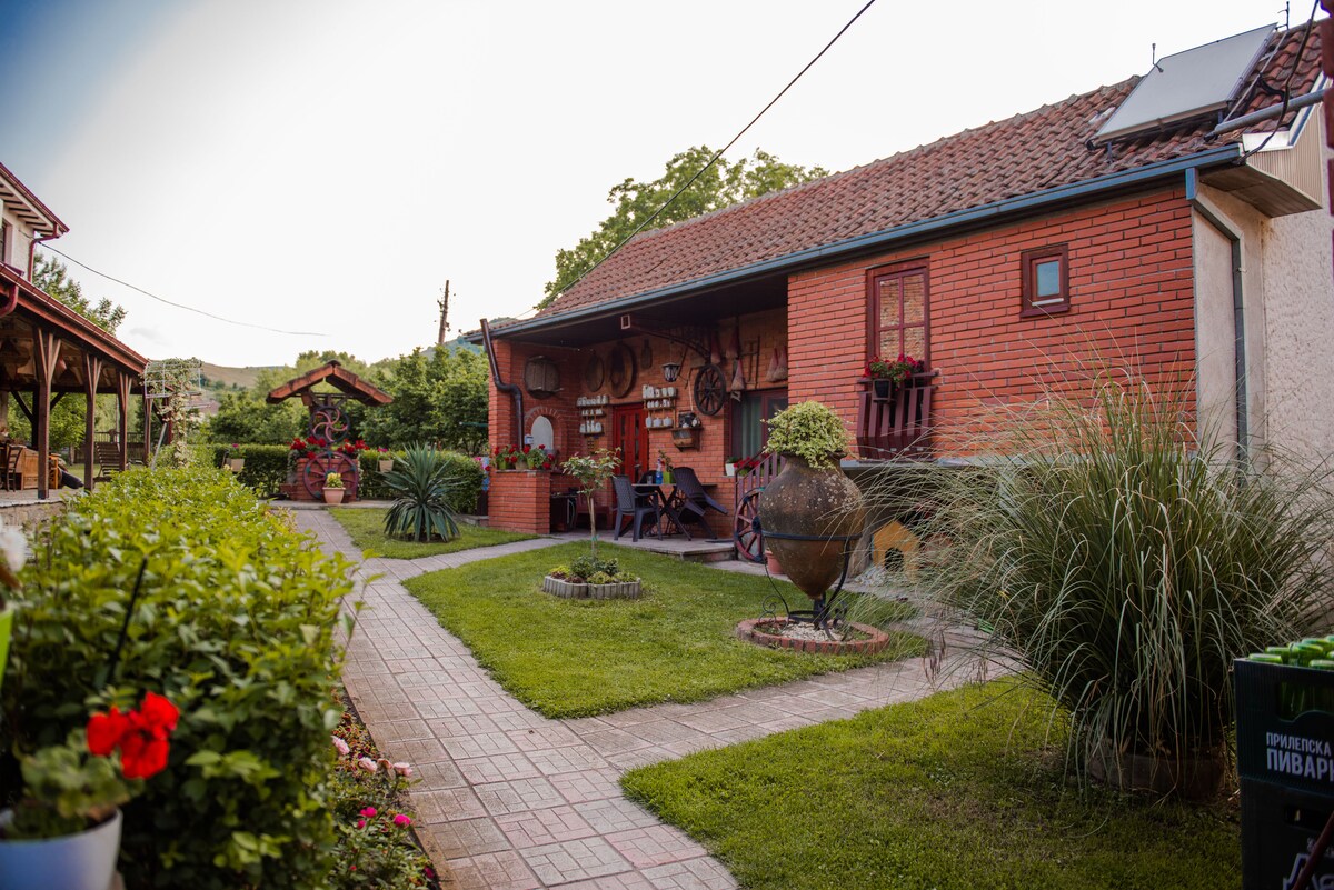 A charming exterior showcases a brick building surrounded by lush greenery and well-maintained pathways. Flowering plants and decorative elements enhance the peaceful setting. The area includes a cozy porch with rustic seating and a barbecue space, inviting relaxation in a tranquil environment.