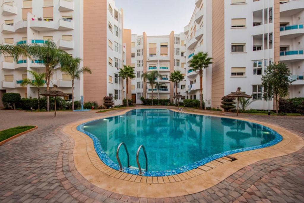 A large swimming pool is surrounded by palm trees and landscaped gardens, located within a residential complex. The pool features a curved shape, with steps leading into the water and seating areas nearby for relaxation.