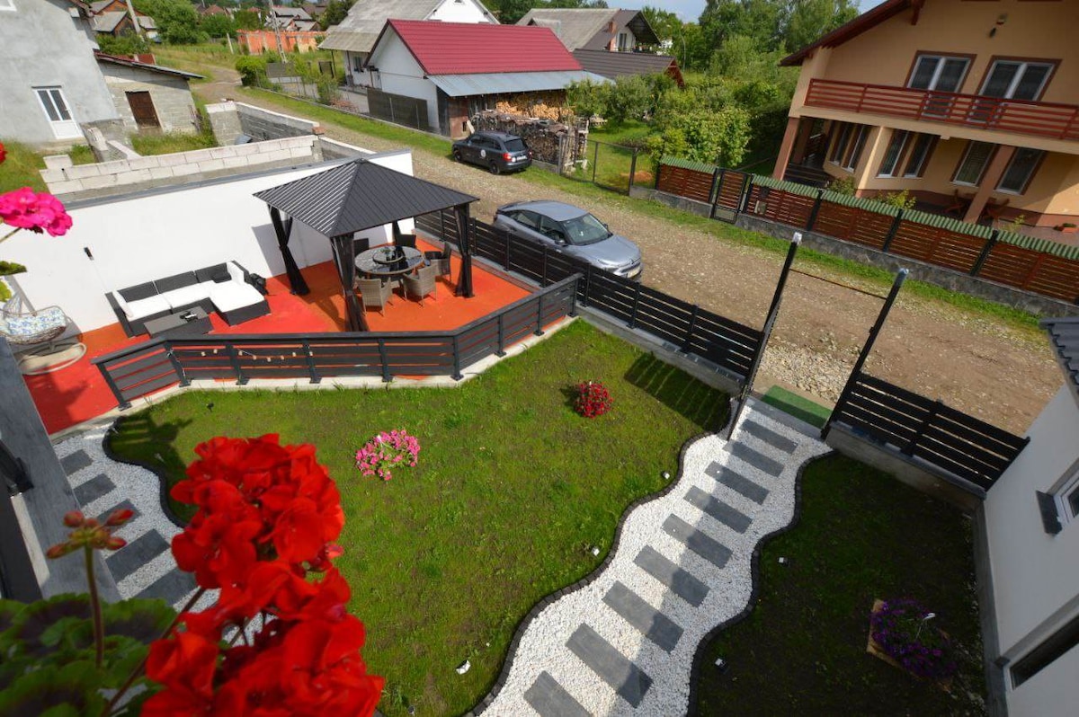 An aerial view displays a well-maintained garden with a vibrant flower bed and a winding stone pathway. A covered outdoor seating area is positioned on a red deck, while a car is parked nearby. The surrounding neighborhood includes various houses and greenery.