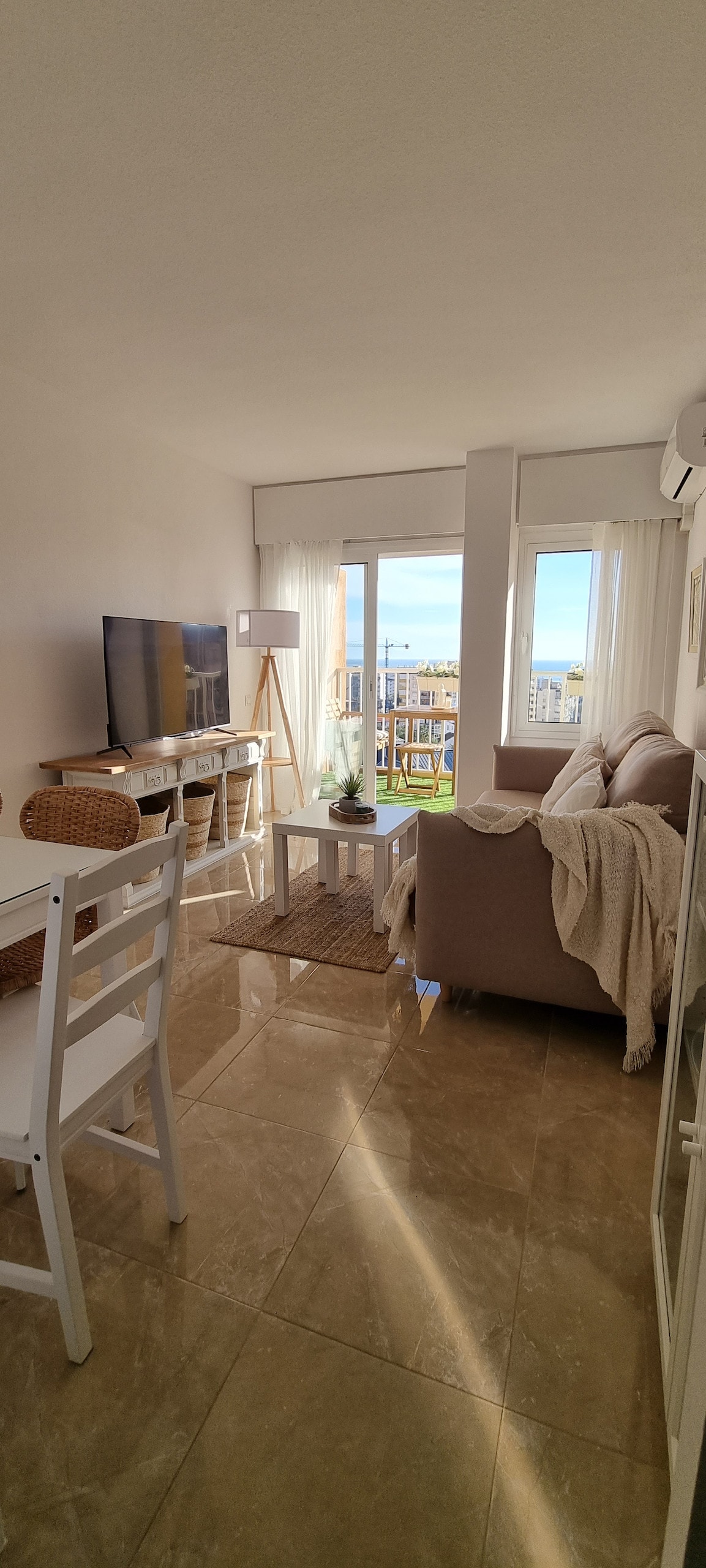 A bright living area presents a comfortable sofa and a television mounted on the wall. Large windows allow natural light to fill the space, providing a view of the outside. A small dining table and chairs are included, along with decorative elements on the shelves.