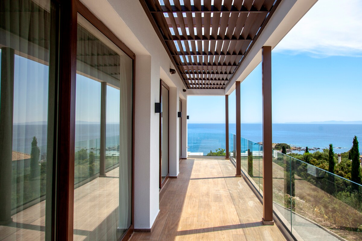A spacious balcony offers panoramic views of the Ionian Sea, framed by large glass panels. Floor-to-ceiling sliding doors provide access to the indoor living areas, while a wooden pergola adds shade. The serene landscape includes greenery and distant shorelines in the background.