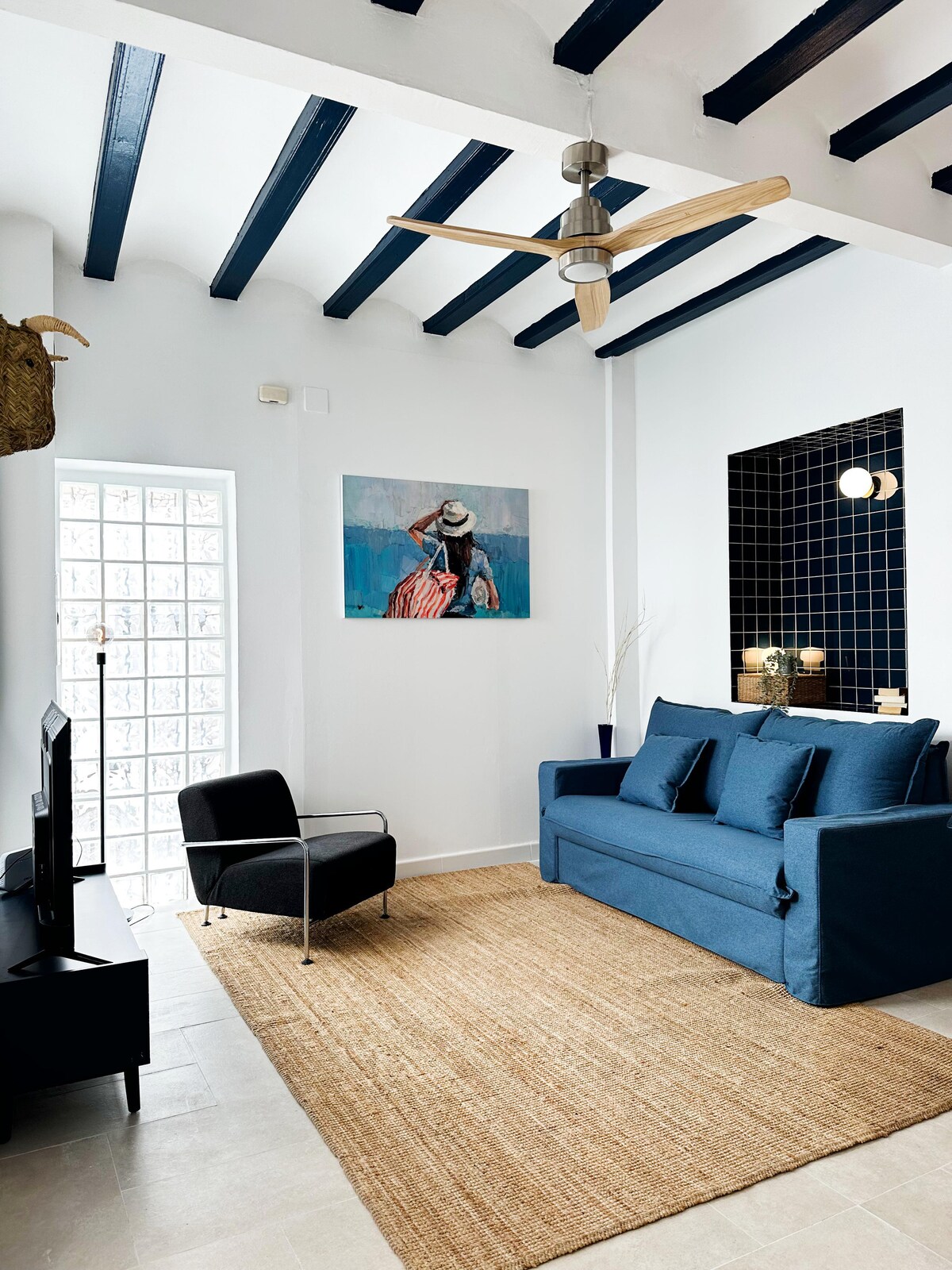 The living area features a blue sofa and a sleek black accent chair, with light streaming in through a frosted glass door. A piece of artwork depicting a woman in a hat adorns the wall, complemented by a woven rug that adds texture to the space.