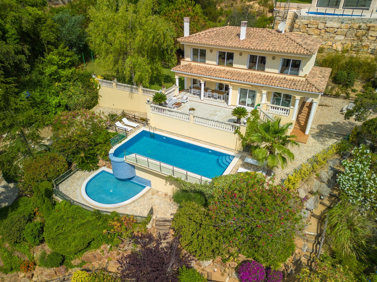 An aerial view of the villa reveals well-maintained gardens and an impressive double pool. The larger pool features a waterfall cascading into a shallower section, perfect for children. Spacious terraces and outdoor dining areas are visible, surrounded by vibrant greenery and flowering plants.
