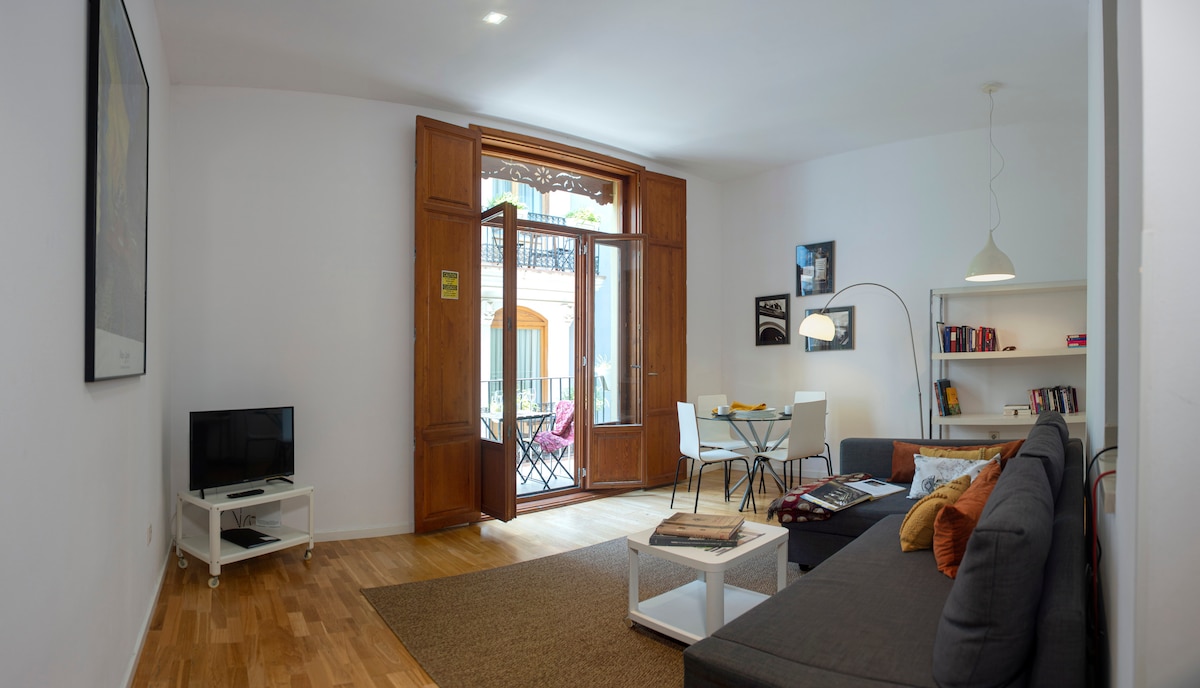 The living area is illuminated by natural light from large wooden doors that open to a balcony. A comfortable gray sofa sits opposite a small dining table, while a bookshelf displays various books. A TV is positioned on a white stand adjacent to the seating area.