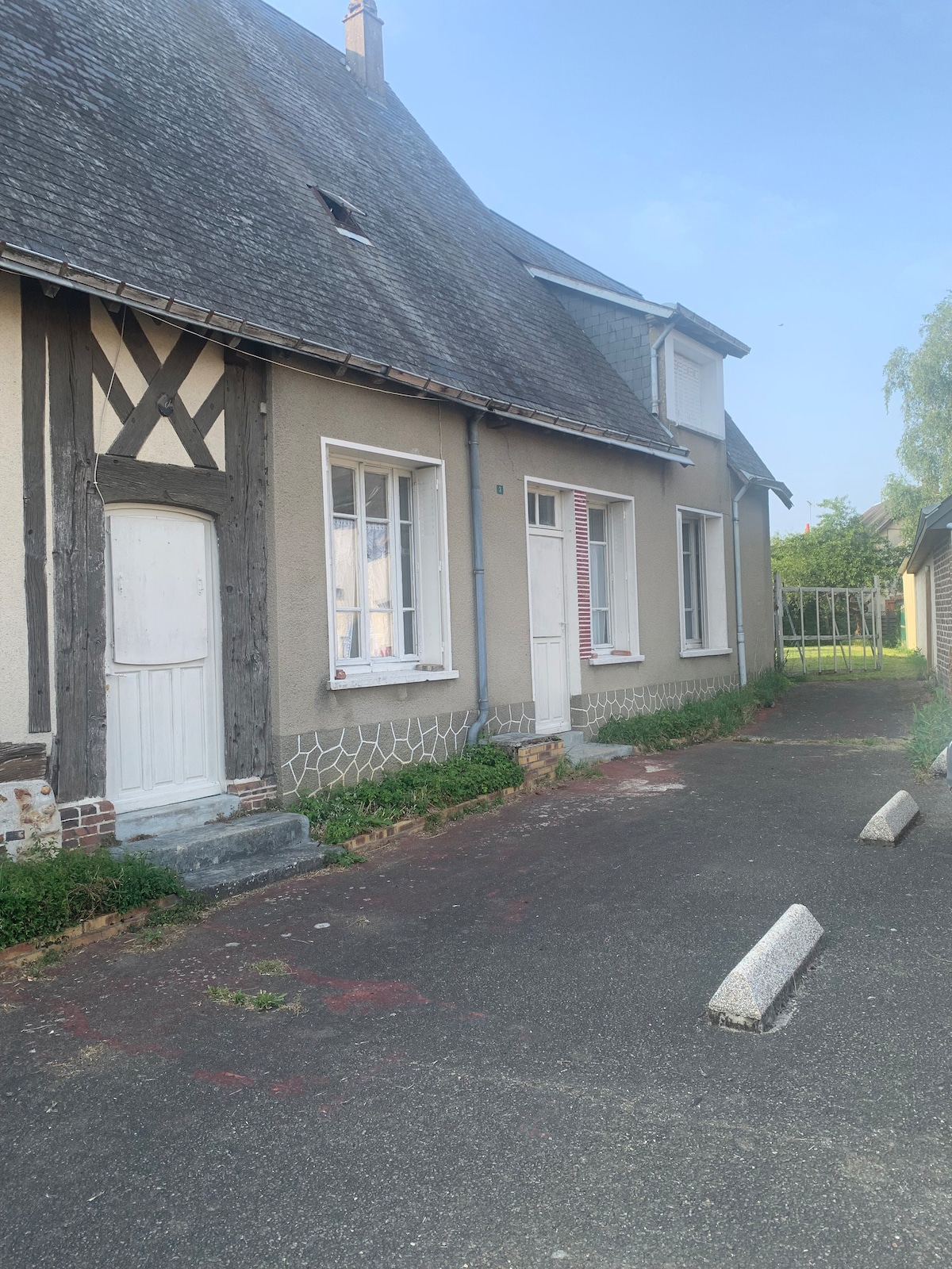 A country-style house is depicted with a slate roof and a combination of plaster and timber-frame siding. The entrance features a white door, with several windows allowing natural light to enter. A gravel path leads to the side garden area, showcasing a peaceful outdoor environment.