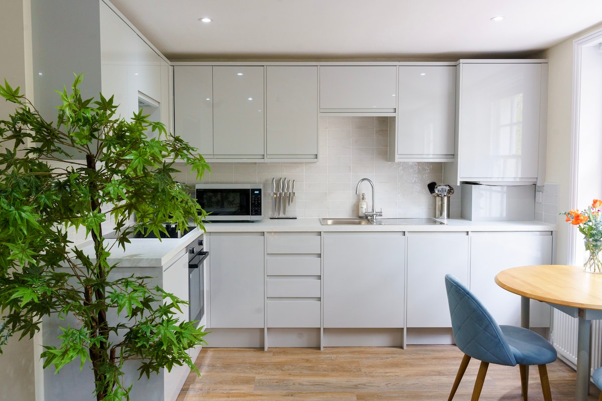 A sleek, modern kitchen features glossy white cabinets and a minimalist design. Stainless steel appliances are integrated into the cabinetry. A small dining table with a round wooden top and a blue chair is positioned nearby, creating a functional space. Natural light brightens the area.