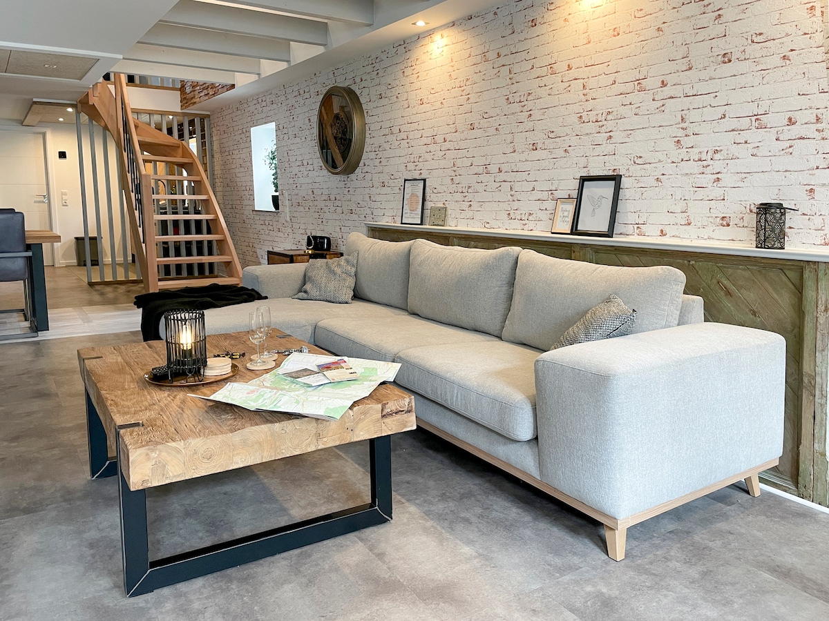 A spacious living area features a light gray sofa, complemented by a wooden coffee table displaying maps and glasses. Exposed brick walls and decorative items accentuate the modern design. Natural light illuminates the space through large windows.