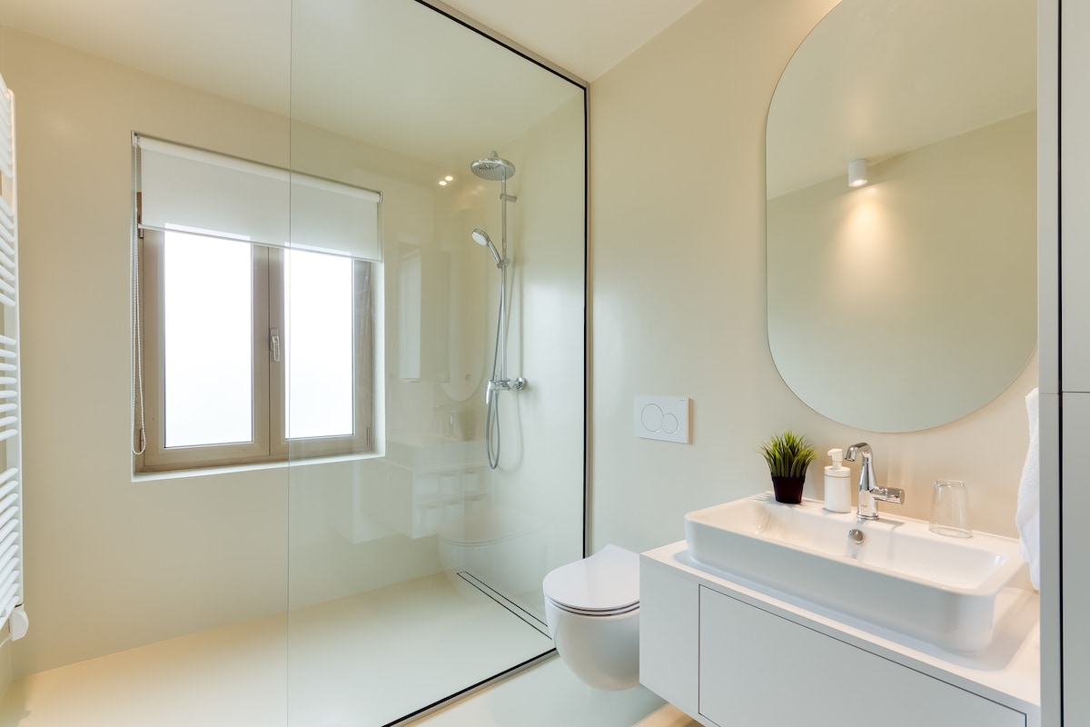 A modern bathroom is featured with a large walk-in shower, complete with a handheld and overhead shower. A sleek sink with a mirror above sits beside a toilet. Natural light enters through a window, while minimalist decor adds to the clean aesthetic.