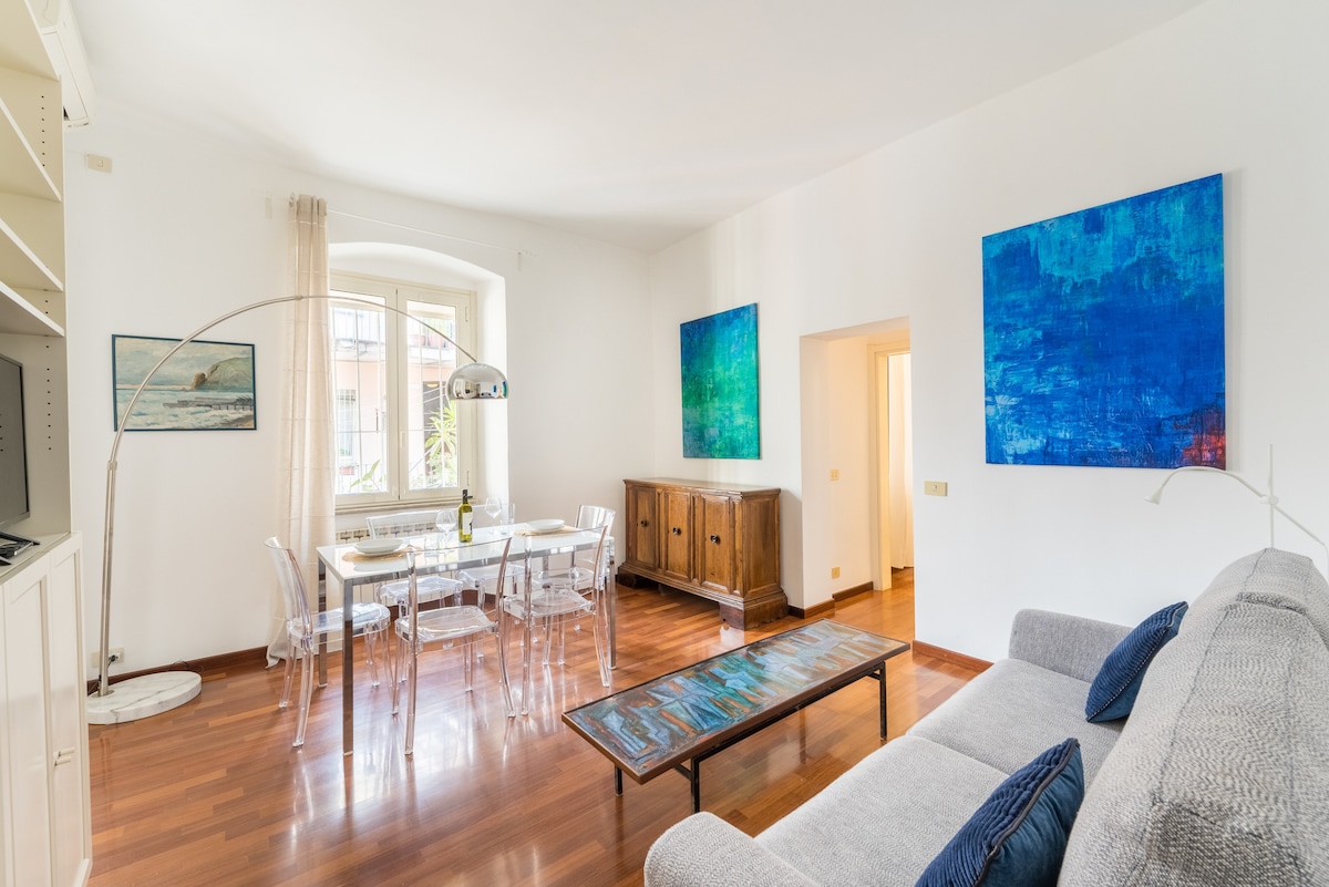 A bright living area features a comfortable gray sofa and a glass coffee table, accented by blue artwork on the walls. A dining table with transparent chairs is situated near a large window, allowing natural light to fill the space.