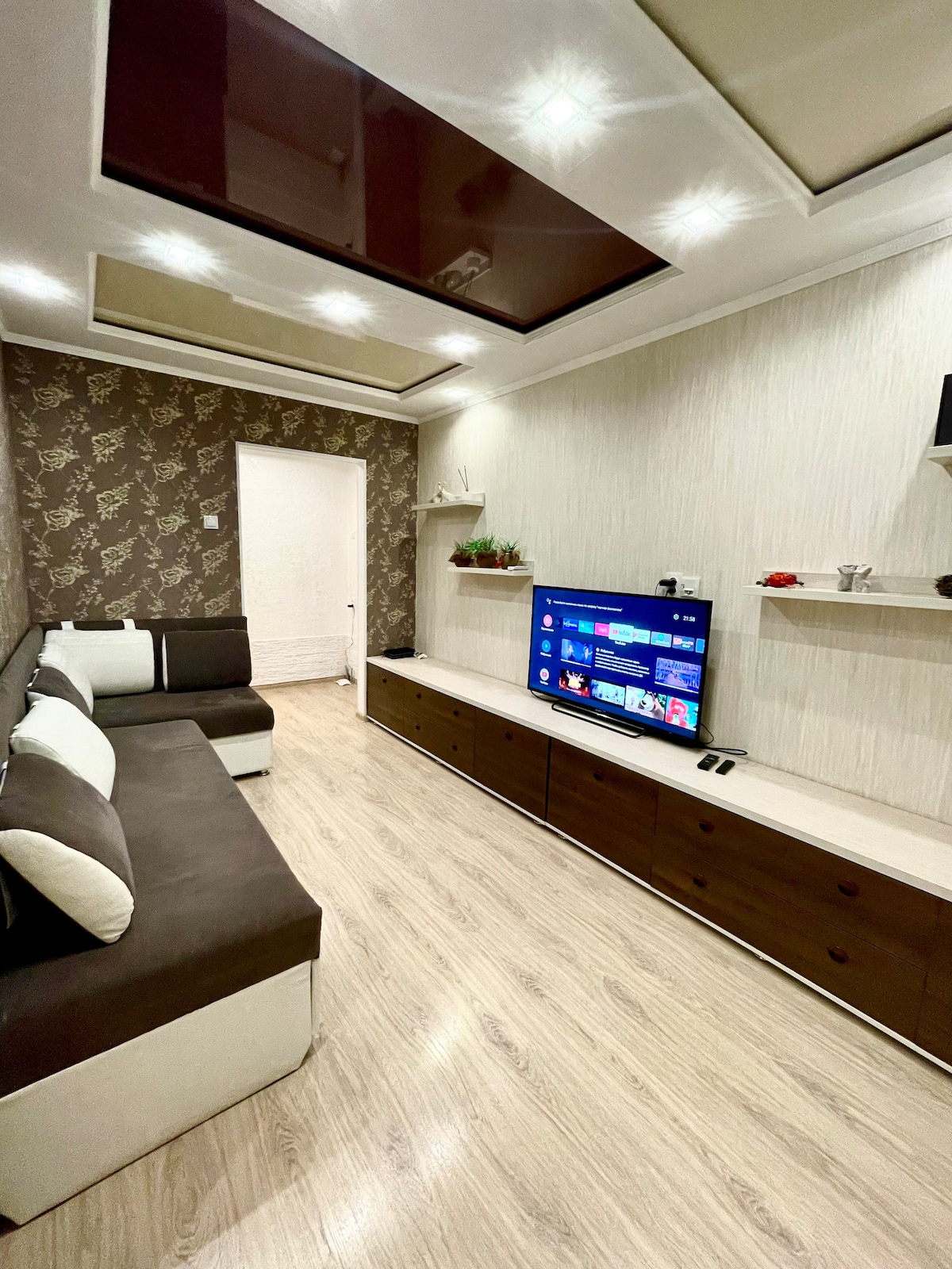 A living area is presented with modern furnishings, featuring a large flat-screen TV mounted on a light wood media unit. A comfortable sectional sofa in contrasting colors offers seating, while decorative shelves add subtle elegance to the wallpapered walls.
