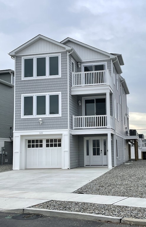 A modern beach house is visible with a gray exterior and multiple large windows. The structure features two balconies and a garage on the ground level. The surrounding driveway is made of concrete, with gravel present in the area, providing a spacious outdoor entry.