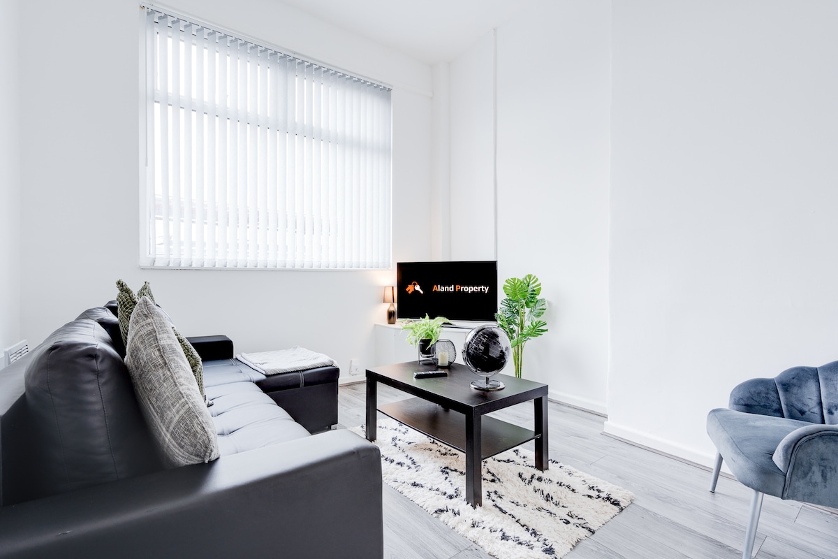 A modern living area features a dark leather sofa accompanied by decorative pillows. A low coffee table sits in the center on a patterned rug. A large screen TV is mounted on the wall, with natural light streaming through horizontal blinds.