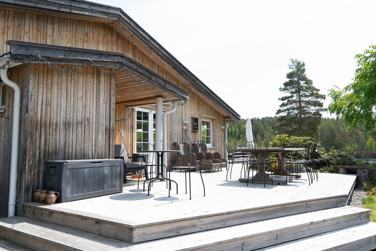 An expansive deck is featured, constructed of wood, and furnished with multiple seating options, including tables and chairs. Lush greenery surrounds the area, promoting a connection to nature. Sunlight bathes the space, creating an inviting outdoor environment for relaxation and socializing.