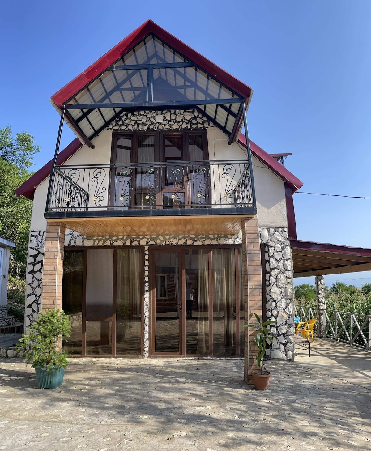 A charming two-story structure is presented, featuring a triangular roof and a balcony adorned with intricate wrought iron details. The entrance is highlighted by clear glass doors, and smooth stone walls complement the surrounding natural landscape. Potted plants and outdoor seating are visible on the patio.