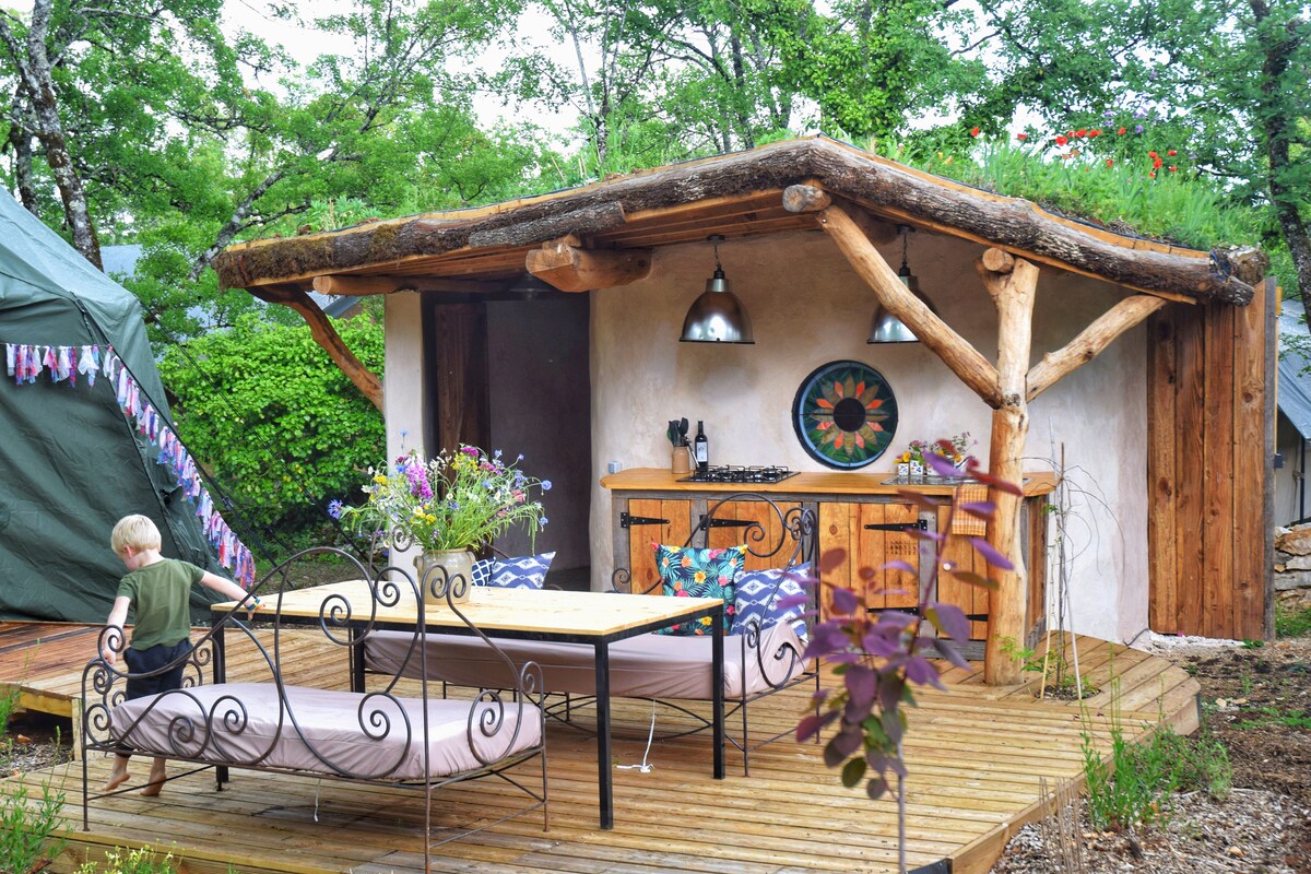 An inviting outdoor kitchen area is situated under a green roof, featuring wooden beams and two hanging light fixtures. A dining table with decorative ironwork chairs is positioned on a wooden deck, accompanied by vibrant potted plants and seating for relaxation.