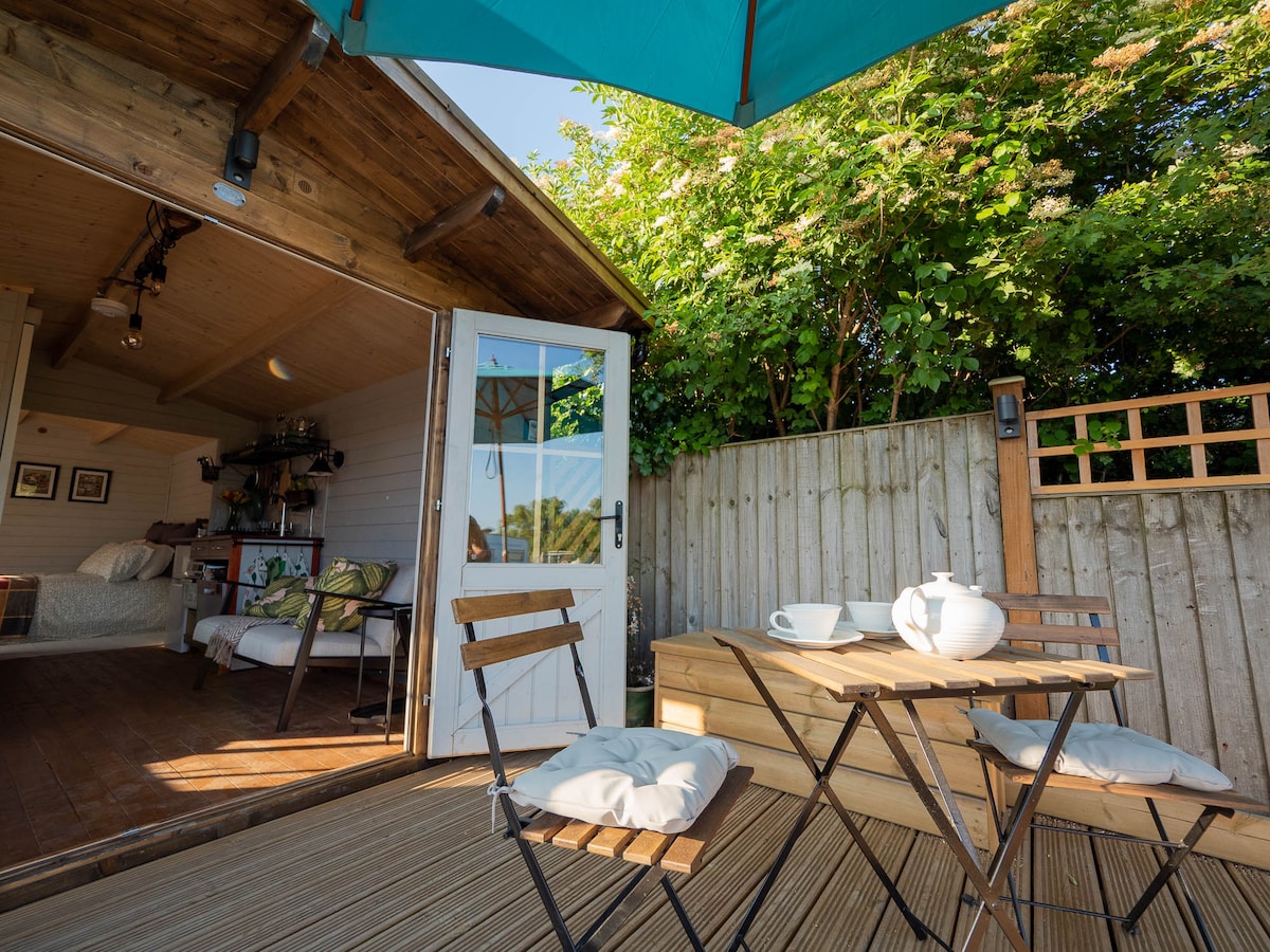 A private outdoor seating area includes a small table with two chairs and a tea set. A large umbrella provides shade. The scene is framed by a wooden fence and lush greenery, creating a serene outdoor space connected to the indoor living area.
