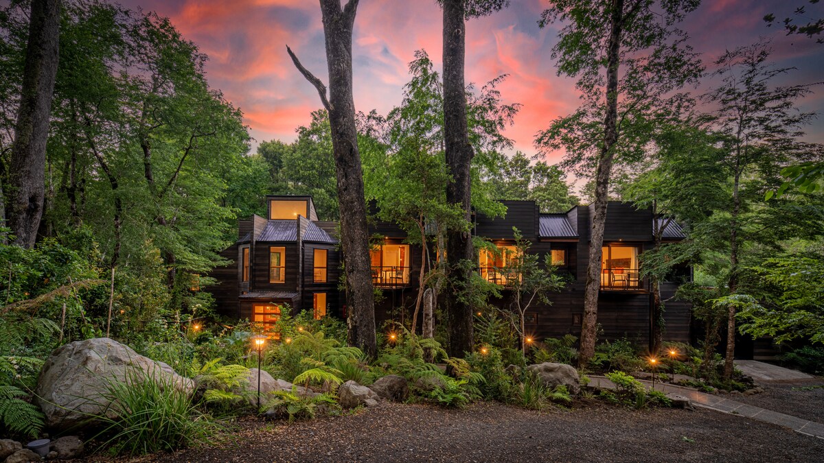 A modern lodge is nestled among towering trees, surrounded by lush greenery and soft lighting illuminating the pathway. The building features multiple balconies framed by large windows, while a colorful sunset sky adds a warm glow to the serene forest setting.