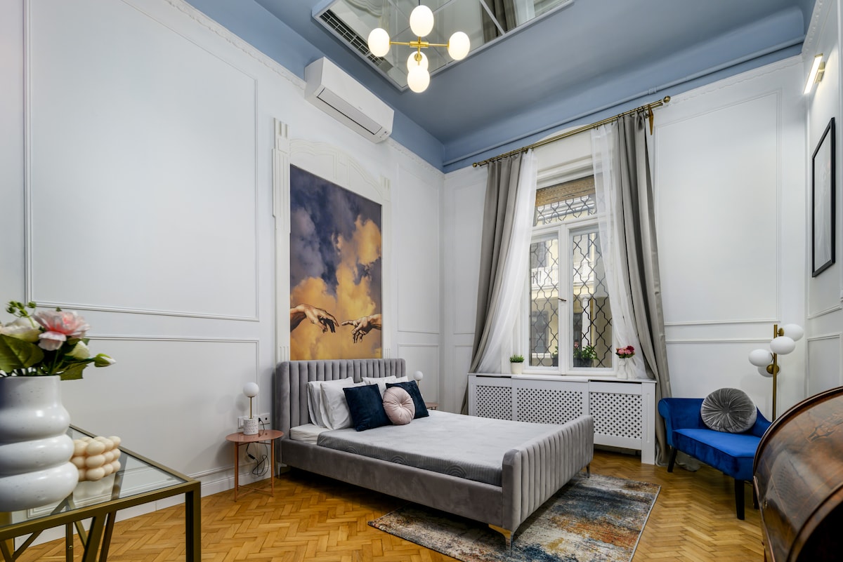 A well-appointed bedroom features a spacious gray bed centered beneath a picturesque wall art piece depicting clouds. Natural light flows through a large window dressed in sheer curtains. A blue accent chair is placed near the window, complemented by stylish decor elements.