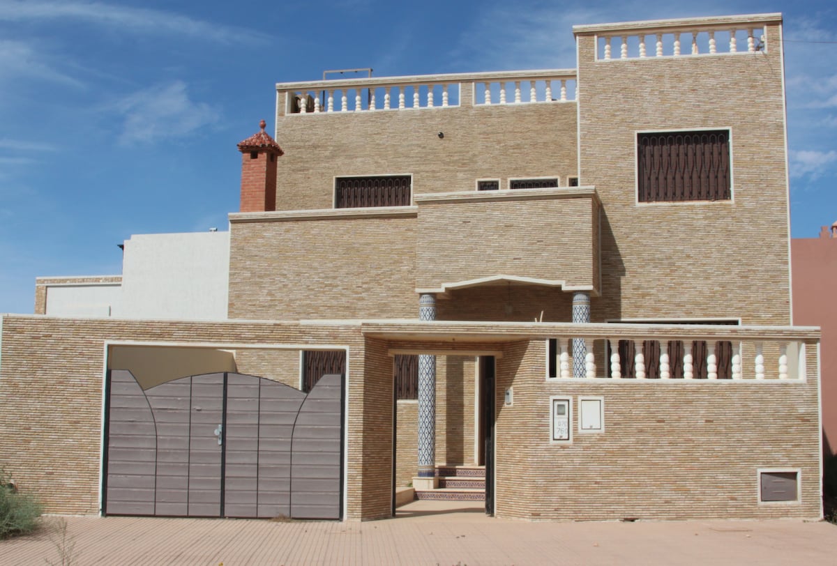 The villa's exterior showcases a unique architectural style with textured stone walls and decorative balcony railings. A large, muted gate leads to the entrance, framed by pathways and shrubs. Clear skies provide a sunny backdrop, illuminating the building's warm colors.