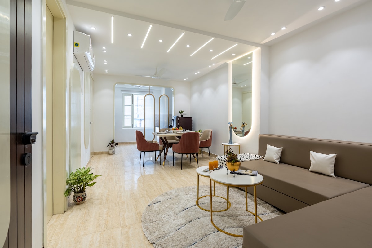 A spacious living area features a modern 5-seater sofa set complemented by two accent chairs and a 4-seater dining table. Soft lighting reflects off the minimalist decor, creating a bright and inviting atmosphere. A round rug softens the space, while potted plants add a touch of greenery.