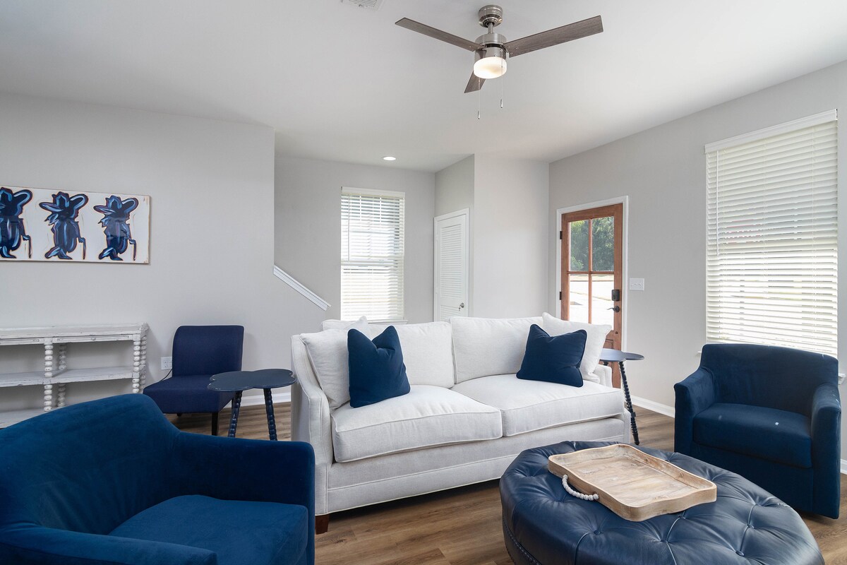 A living area features a white sofa with navy blue accent pillows, positioned near large windows allowing natural light. Two blue armchairs complement a round ottoman with a wooden tray on top. A light ceiling fan circulates air in the spacious room.