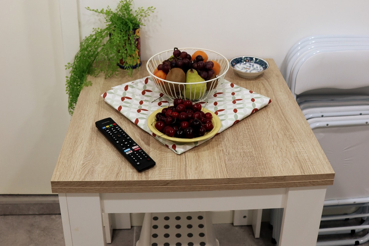 A wooden table is set with a bowl of assorted fruits, including grapes and cherries, and a small decorative dish. A remote control is placed beside a folded patterned cloth. A potted plant adds a touch of greenery in the background.