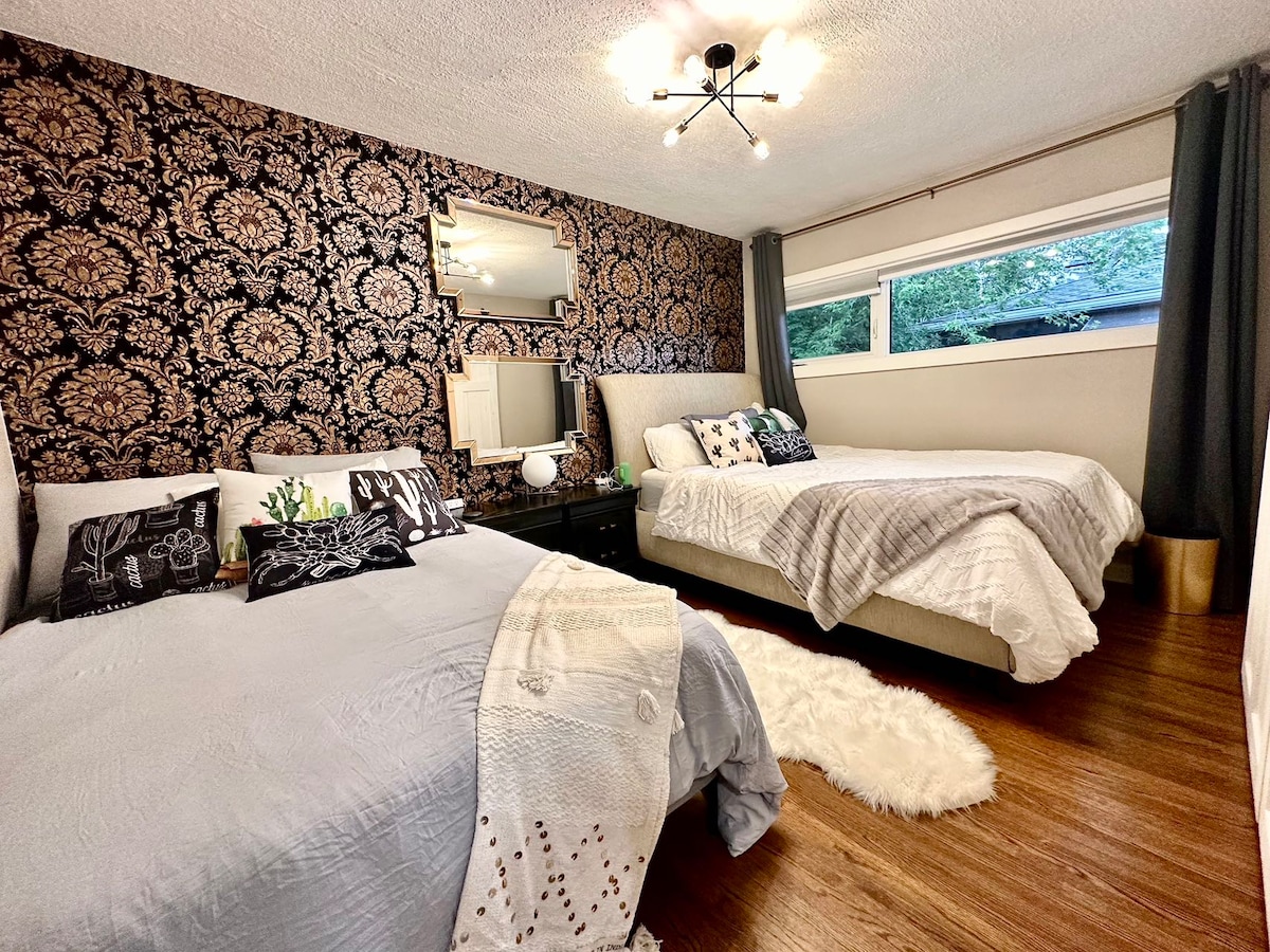 A cozy bedroom is presented with two beds: one with light grey bedding and the other with a light cover. Elegant dark floral wallpaper enhances the space, while a mirror reflects light from the window above. Accents include decorative pillows and a plush rug near the floor.