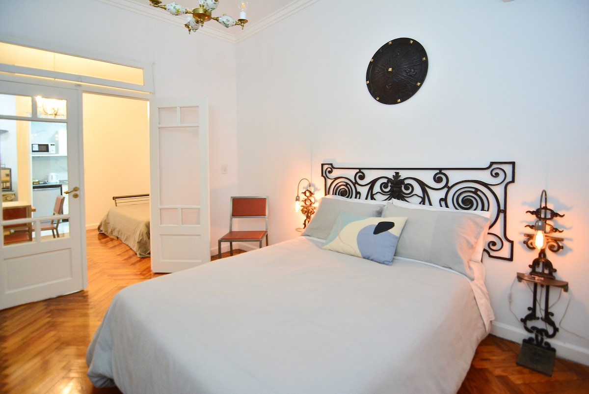 A spacious bedroom features a large bed with a light grey coverlet, complemented by decorative wall sconces on either side. A soft pillow is placed on the bed. The room also includes a glimpse of a second bed and a doorway leading to a kitchen area.