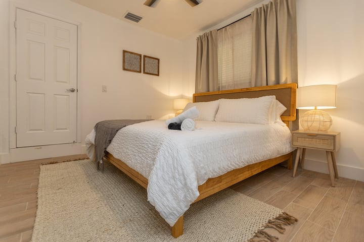 Chumba cha kulala cha mtindo wa boho-chic katika Casa Bali huko Miami chenye kitambaa cheupe cha kitanda, karatasi ya ukutani yenye ruwaza isiyo na upande wowote, taa za ukutani za kijiti zenye mwanga wa joto na meza za kuweka taa za usiku zilizofumwa.