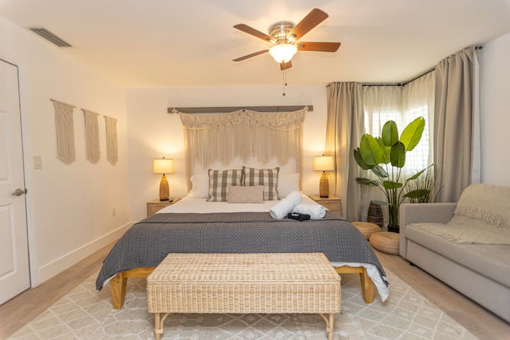 Chumba cha kulala cha mtindo wa boho katika Casa Bali huko Miami kilicho na kitanda chenye blanketi la kijivu, kichwa cha kitanda cha kipekee cha macrame, benchi ya rattan iliyofumwa kwenye sehemu ya miguu na taa za joto za kando ya kitanda karibu na mmea mkubwa wa kitropiki.