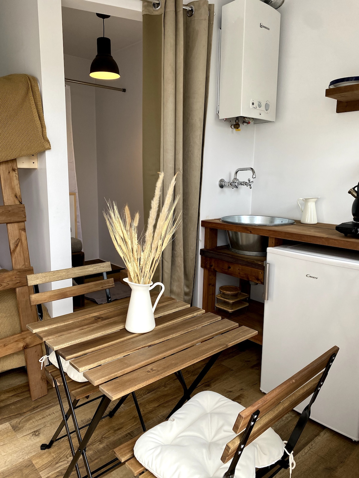 An inviting kitchenette area features a small wooden table with two folding chairs. A simple vase with dried grasses sits on the table. The compact space includes a sink, a white refrigerator, and wooden shelves, contributing to a cozy atmosphere.